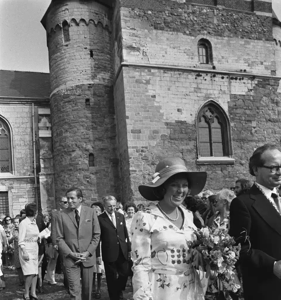 Photographie en noir et blanc : la princesse Beatrix des Pays-Bas, un bouquet à la main, entourée d’officiels devant une église